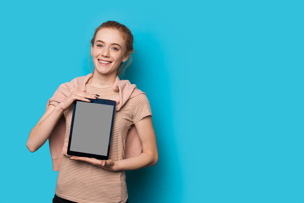 Caucasian woman with red hair and freckles showing a tablet with blank space on blue wall with free space - Photo, Image