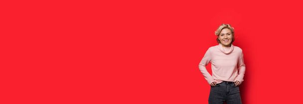 Mulher caucasiana encantadora posando em uma parede vermelha com espaço livre de mãos dadas no bolso
 - Foto, Imagem