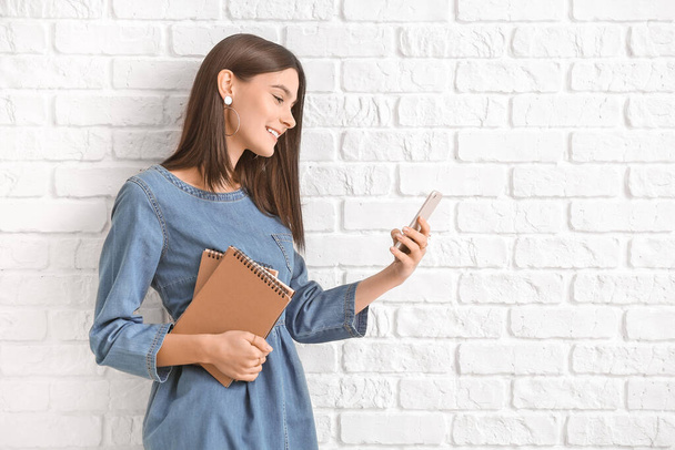 Beautiful young woman with mobile phone on white brick background - Zdjęcie, obraz