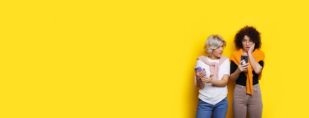 Caucasiano encaracolado menina olhando chocado em seu celular enquanto posando perto de seu amigo em uma parede amarela com espaço livre
 - Foto, Imagem