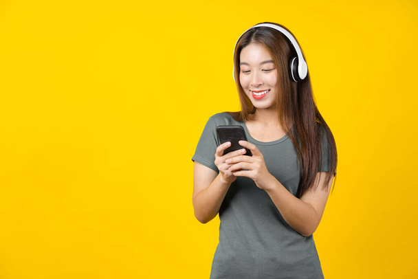 Happiness Asian uśmiechnięta młoda kobieta noszenie technologii bezprzewodowe słuchawki do słuchania muzyki za pośrednictwem inteligentnego telefonu komórkowego na izolowanym tle żółty kolor, Styl życia i wypoczynek z koncepcji hobby - Zdjęcie, obraz