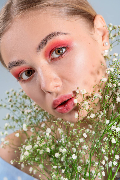 Νεαρή γυναίκα με όμορφο μακιγιάζ και λουλούδια σε γκρι φόντο - Φωτογραφία, εικόνα