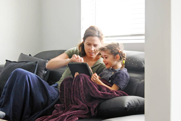Mãe e filha usando um tablet na sala de estar, enquanto forçado a ficar em casa, porque as políticas do governo esforços para evitar a propagação do coronavírus pandêmico (COVID-19). Pessoas reais. Espaço de cópia
. - Foto, Imagem