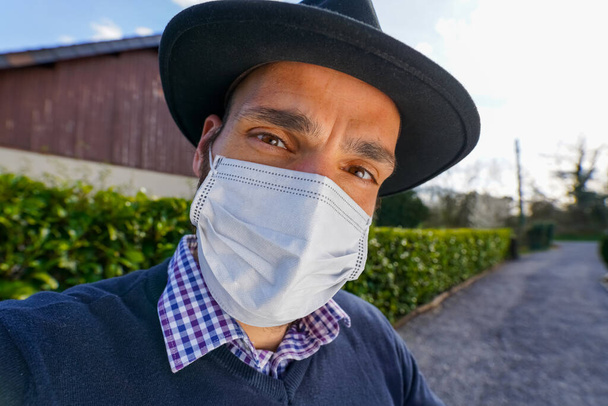 Homem usando máscara branca e chapéu preto em casa durante o confinamento e quarentena. Coronavirus Covid-19, SARS-CoV-2
 - Foto, Imagem