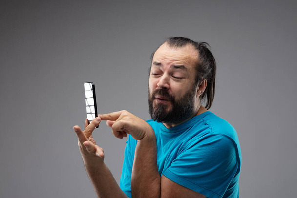 Brodaty mężczyzna w niebieskiej koszulce szturchający ekran telefonu z delikatnym grymasem na twarzy. Portret z bliska na szarym tle z przestrzenią do kopiowania - Zdjęcie, obraz