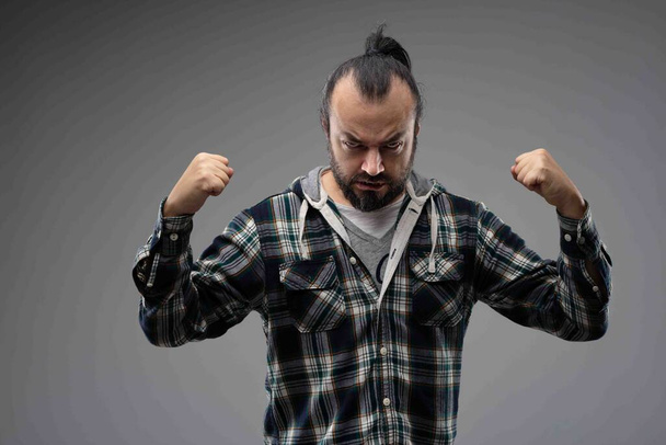 Γενειοφόρος άνδρας με χτενισμένα μαλλιά, στέκεται σε καρό πουκάμισο με σφιγμένες γροθιές, σαν να δείχνει τη δύναμη και το θυμό του πριν από τον αγώνα. Εμπρόσθιο πορτρέτο σε γκρι φόντο - Φωτογραφία, εικόνα