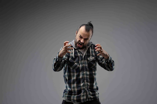 Szakállas férfi fésült fekete hajjal, ellenőrzött ingben, agresszív grimaszokkal és feltartott kezekkel. Félhosszú elülső portré stúdióban szürke háttérrel - Fotó, kép