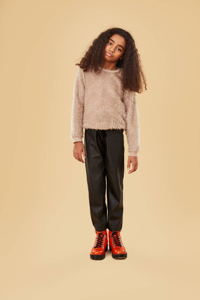 Full body African American child in trendy outfit tilting head and looking at camera while standing against beige background - Foto, Imagen