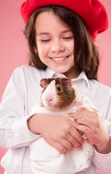 Cute happy little girl in white shirt and red beret holding guinea pig wrapped in white cloth while standing against pink background - Фото, зображення