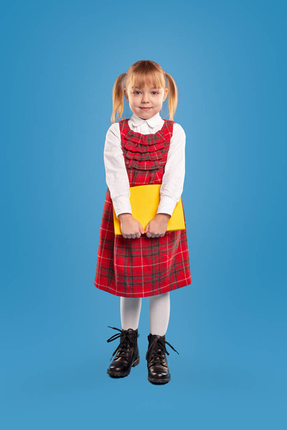 Full body cute girl in school uniform carrying textbook and looking at camera during lesson against blue background - Foto, Bild