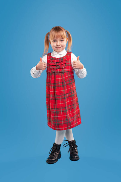 Cute little girl in red checkered sundress and white elegant shirt and black shoes smiling at camera and giving like, while standing alone against blue background - Foto, immagini