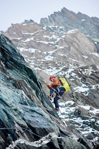 Widok z boku człowieka wspinaczka z plecakiem za pomocą stałej liny, aby wspiąć się na wysoką skalistą górę, wspinaczka grzbiet alpejski i próbuje dotrzeć do szczytu góry. Koncepcja wspinaczki górskiej i alpejskiej. - Zdjęcie, obraz