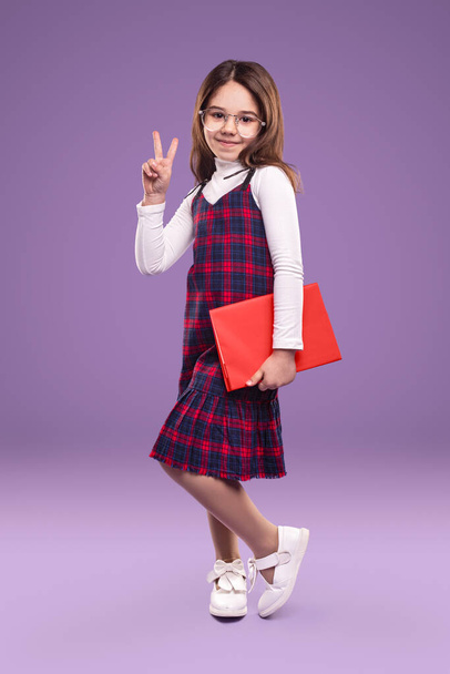 Full body adorable child in school uniform carrying textbook and gesturing V sign against violet background - Photo, image