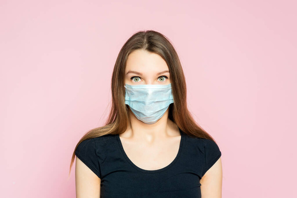 Coronavirus, masked woman.Studio portrait of a young woman wearing a face mask, looking at camera, on pink background. - Фото, изображение