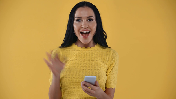 Mujer sonriente con teléfono inteligente agitando la mano en la cámara aislada en amarillo
 - Imágenes, Vídeo