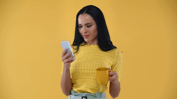 Χαμογελαστό κορίτσι με κύπελλο χρησιμοποιώντας smartphone και κοιτάζοντας κάμερα απομονωμένη στο κίτρινο - Πλάνα, βίντεο