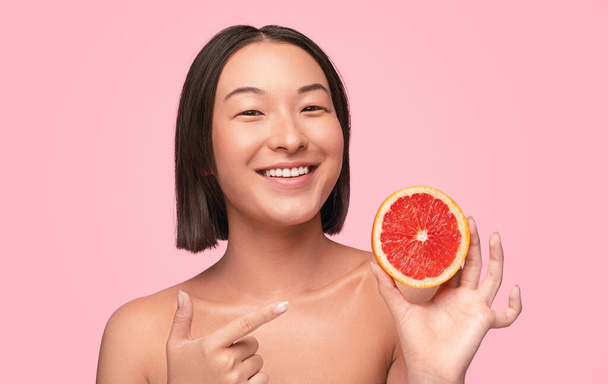 Happy healthy young Asian female with bare shoulders demonstrating sliced ripe red grapefruit on pink background - Photo, Image