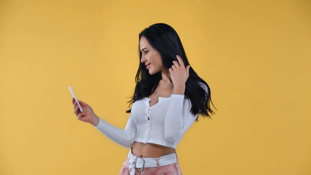 Smiling woman taking selfie with smartphone isolated on yellow - Metraje, vídeo