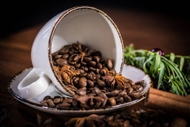An upturned Cup with scattered coffee beans, decorated with cinnamon sticks, tarragon sprigs and cherries. - Photo, Image