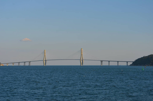 Eine Brücke spannt sich über eine Bucht von einer Insel zum Festland. Zwei Pylonen mit stützenden Metallstreben ragen über die Brücke und bilden ein architektonisches Merkmal. Der Himmel ist blau mit Wolken. - Foto, Bild