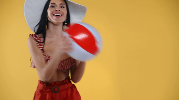 Uśmiechnięta dziewczyna w kapeluszu rzuca nadmuchiwaną piłkę izolowaną na żółto - Materiał filmowy, wideo