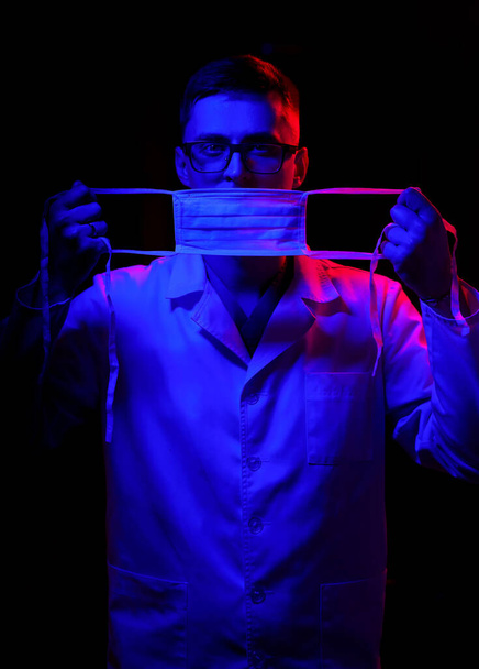 Portrait of doctor in scrubs. Black background with blue - red light. Doctor in glasses closed mouth with medical mask. Close up shot. - Photo, image