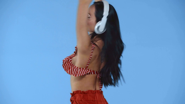 Chica sonriente en auriculares sosteniendo teléfono inteligente y bailando aislado en azul
 - Metraje, vídeo