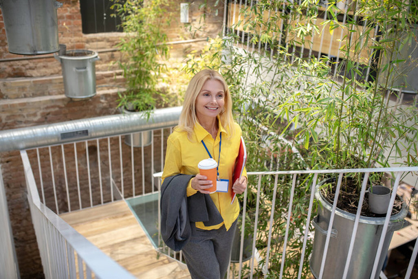 Blonde woman climbing up stairs, holding cup of coffee and jacket, smiling - Photo, Image