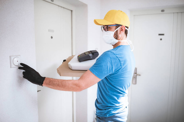 Deliveryman with protective medical mask holding pizza box and POS wireless terminal for card paying, ringing at the doorbell - days of viruses and pandemic, food delivery to your home and safety hygiene measures. - Photo, Image
