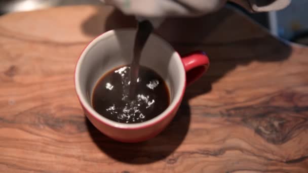 Close-up image of a red coffee cup on the outside and white on the inside resting on a wooden cutting board. A drizzle of steam rises from the cup. Blurry background. - Footage, Video