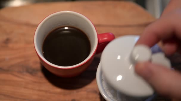 Close up shot of a red coffee cup on the outside and white on the inside resting on a wooden cutting board. A man's hand takes off the lid of the sugar bowl and puts a teaspoon of sugar in the coffee. - Footage, Video