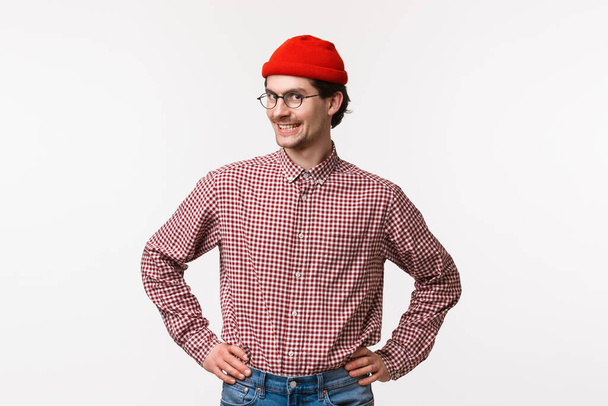 Талия портрет оптимистичный и восторженный молодой хипстер парень в красной шапочке и очках, в клетчатой рубашке, стоя готовый с уверенной прямой позе, держаться за руки на талии и улыбающаяся камера
 - Фото, изображение