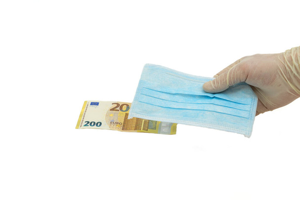 A hand in protective gloves holds out a blue medical mask for money of 200 euros. Concept of buying a mask in Europe, protection against the virus. Isolated white background. - Foto, afbeelding