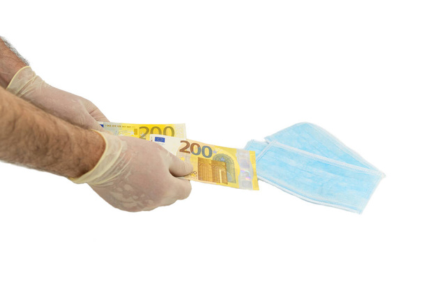Two hands in white protective gloves buy a pack of blue medical masks for money with 200 Euro banknotes. Concept of buying a mask in Europe, protection against the virus. Shortage of medical masks, high cost. Isolated white background. - Foto, imagen