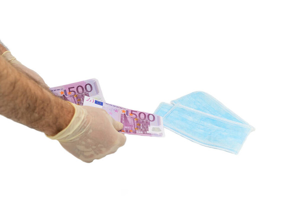 Two hands in white protective gloves buy a pack of blue medical masks for money with 500 Euro banknotes. Concept of buying a mask in Europe, protection against the virus. Shortage of medical masks, high cost. Isolated white background. - Photo, image