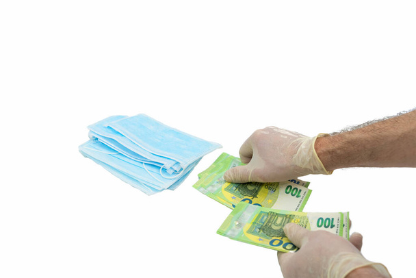 Two hands in white protective gloves buy a pack of blue medical masks for money with 100 Euro banknotes. Concept of buying a mask in Europe, protection against the virus. Shortage of medical masks, high cost. Isolated white background. - Photo, image