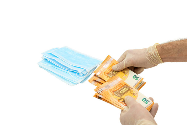 Two hands in white protective gloves buy a pack of blue medical masks for money with 50 Euro banknotes. Concept of buying a mask in Europe, protection against the virus. Shortage of medical masks, high cost. Isolated white background - Photo, Image