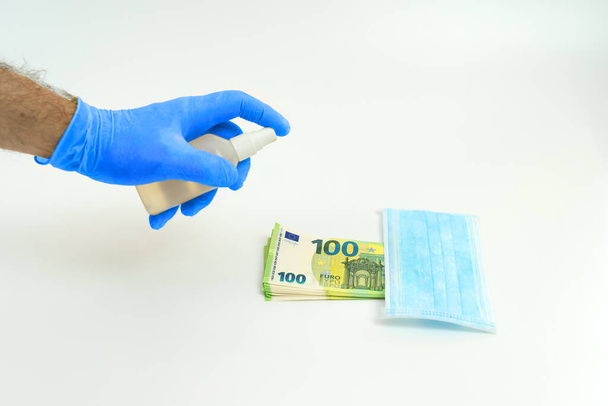 A hand in a blue glove handles with an antiseptic a banknote of money one hundred and 100 euros, next to a blue medical mask. The concept of virus protection is the use of precautions when shopping in stores in Europe. - Photo, Image