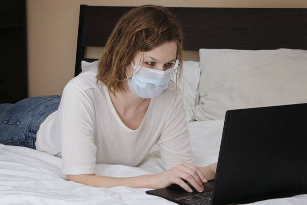 Woman wearing medical mask working lying on a bed with laptop computer. Corona virus quarantine, isolation period covid - 19. Social distancing - Photo, image