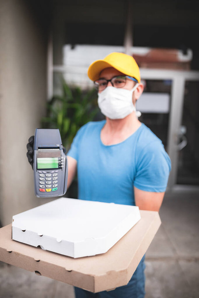 Deliveryman with protective medical mask holding pizza box and POS wireless terminal for card paying - days of viruses and pandemic, food delivery to your home and safety hygiene measures. - Foto, imagen