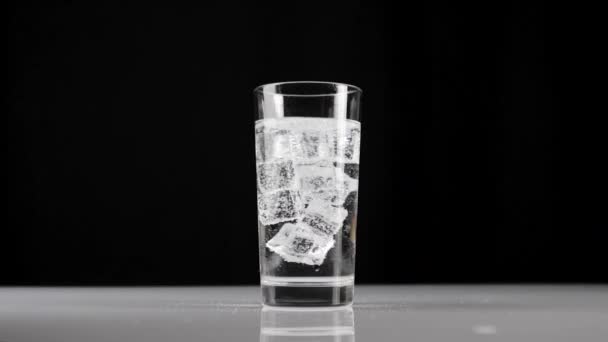 Os cubos de gelo giram e flutuam em um copo com água mineral com bolhas ou refrigerante contra fundo preto. Conceito de ficar em casa e comer alimentos saudáveis com água natural. A meio. Movimento lento
 - Filmagem, Vídeo