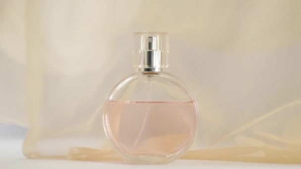 Oválná lahvička s růžovými parfémy nebo esenciálními oleji je na bílém stole. Béžová tkanina se třepotá a mává ve vzduchu kolem láhve. Pojem aroma a vůně. Zavřít. Zpomalený pohyb - Záběry, video