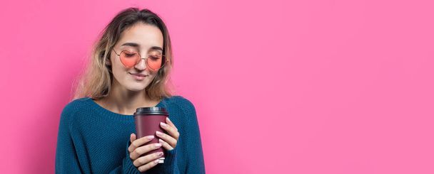 Mujer glamour en gafas en un suéter azul con una bebida de café sobre un fondo rosa
 - Foto, imagen