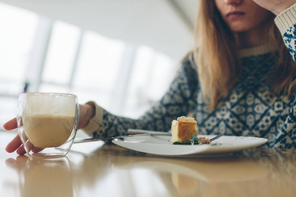 Closeup of glass with double bottom with coffee and peace of dessert and leaf of mint on white plate. Cut view of blonde girl. She wants to take cup. Blurred background. - Foto, imagen