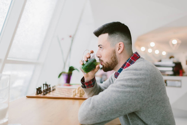 Young bearded man in cafe. He dressed in gray pullover and plaid shirt. Sits at long table near window. Man drinks beverage with green leaves. On table chessboard. Laconic interior. - Photo, Image