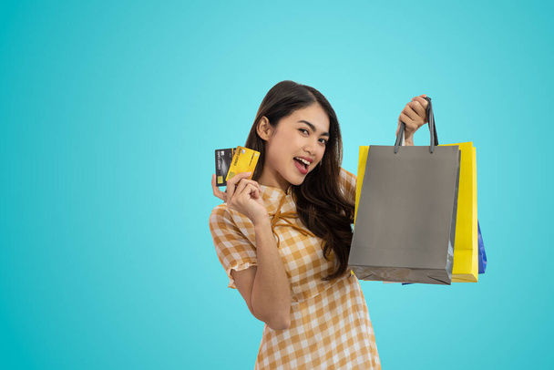 Young happy Asian woman holding shopping bags and showing credit card isolated on blue background - Foto, afbeelding