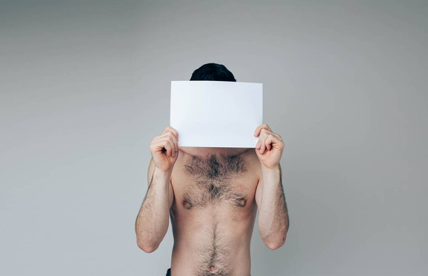 Young man isolated over background. Naked ordinary guy cover face with white blank page. Hide form somebody. Posing on camera - Foto, afbeelding