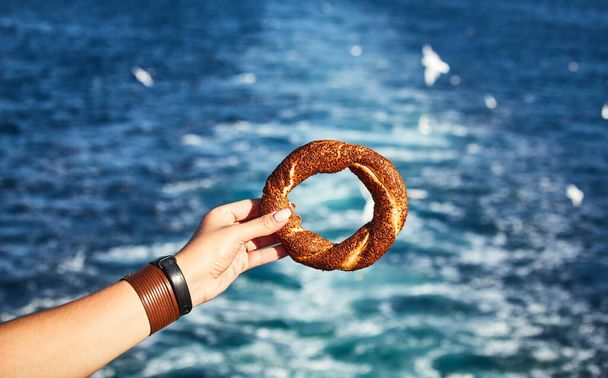 Simitci- um tradicional bagel turco redondo com sementes de sésamo na mão no fundo do Mar de Mármara e na paisagem urbana de Istambul
 - Foto, Imagem