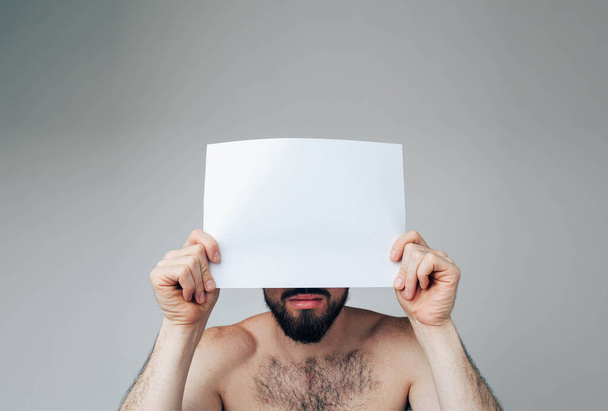 Young man isolated over background. Portrait of male person or model cover face with white blank page. Low part of face is not hidden. Brunette pose on camera. - Photo, Image