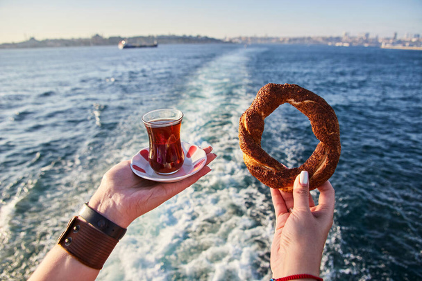A cup (armud) of traditional Turkish tea and Simitci (a round bagel with sesame seeds) in hand against the background of the cityscape of Istanbul and the Sea of Marmar - Photo, Image
